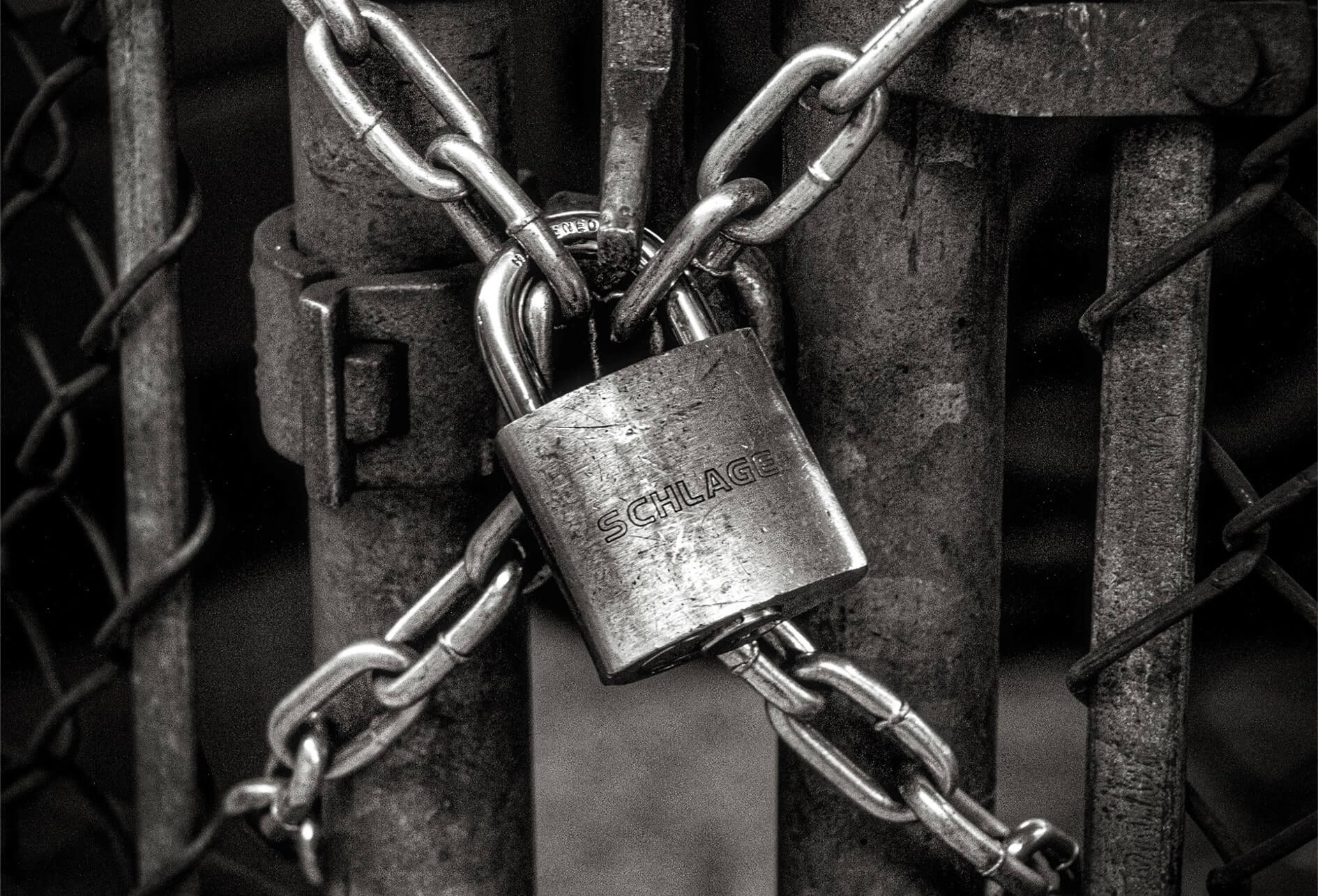 En noir et blanc, un portail grillagé verrouillé par un cadenas et des chaînes.