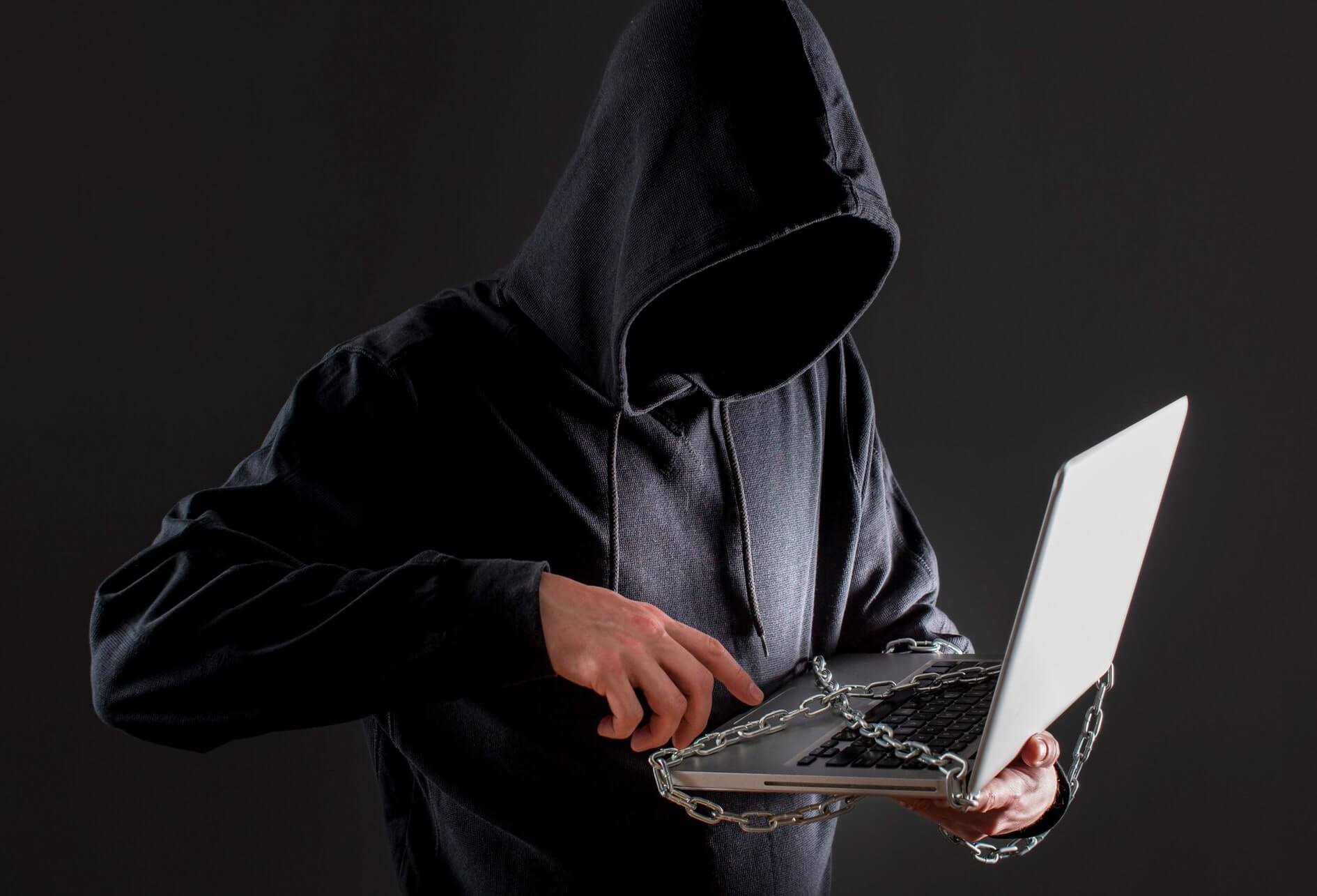 Sur fond sombre, un pirate avec capuche manipule un ordinateur couvert de chaînes.